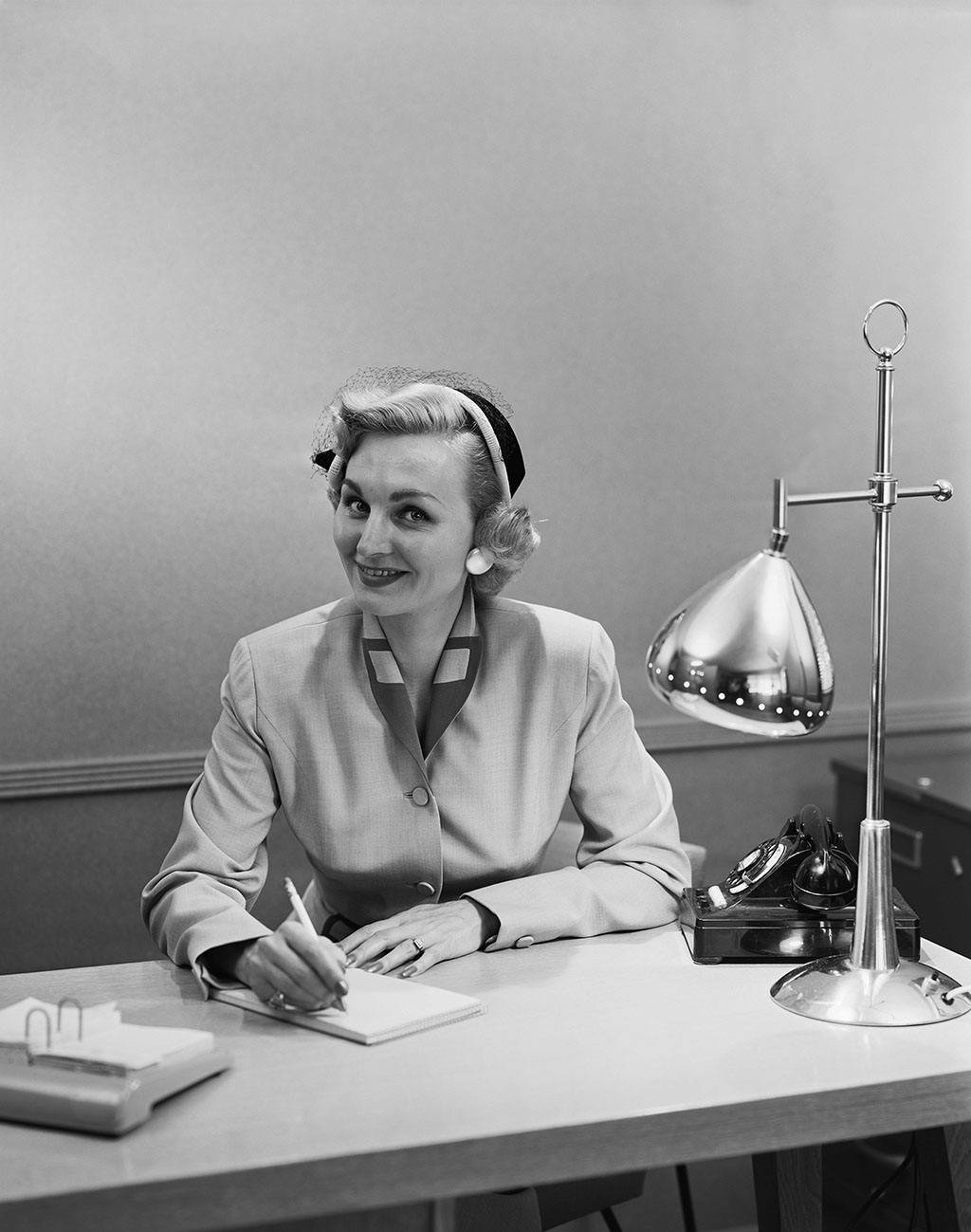 A woman in 1950s attire sits at a desk A woman in 1950s attire sits at a desk, smiling as she writes on a notepad. The desk holds a lamp, a rotary phone, papers, and a pen holder. She wears a suit jacket and a hat with a small veil.