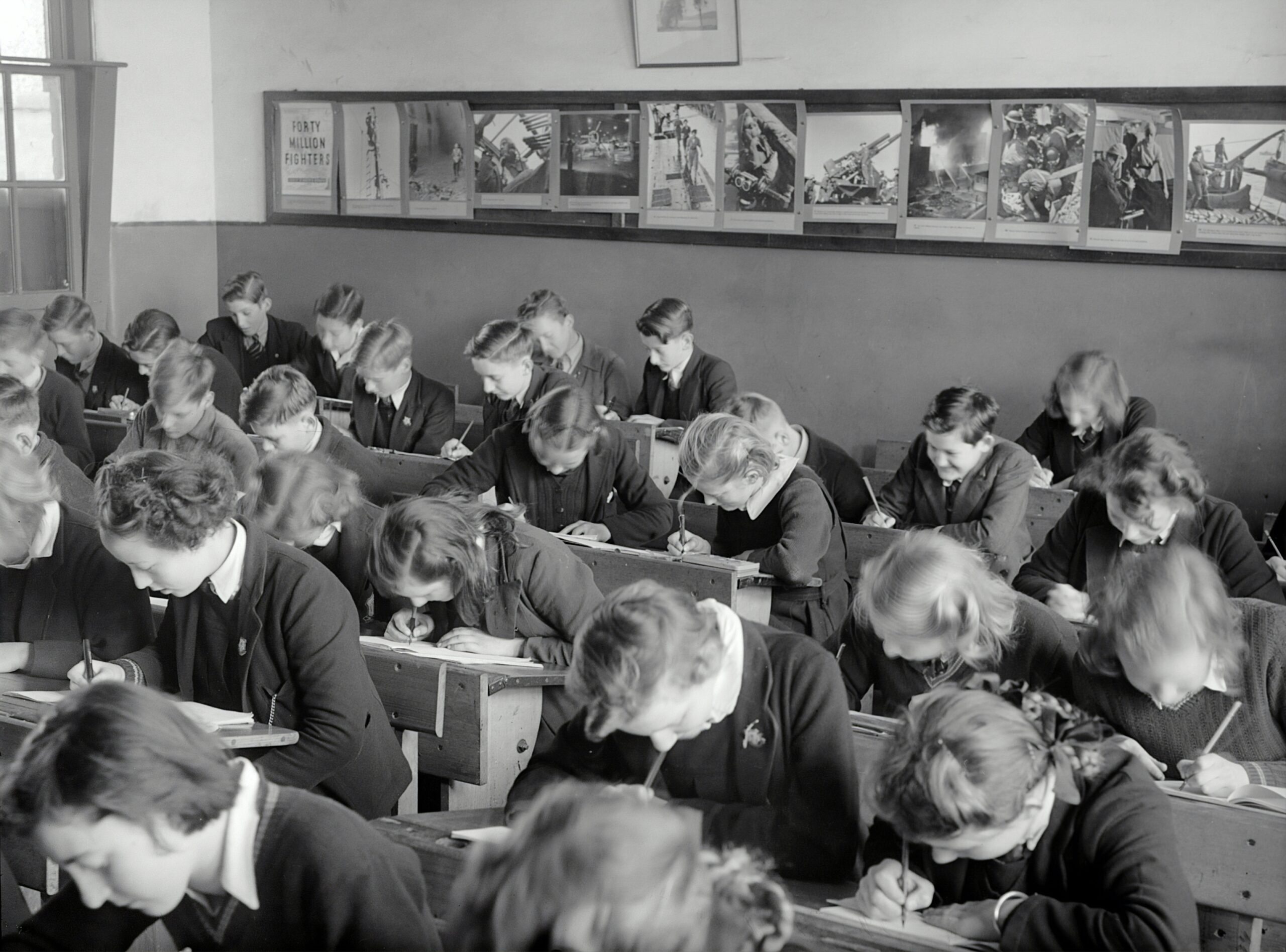 A black and white photo of a classroom filled with students in uniform A black and white photo of a classroom filled with students in uniform, sitting at wooden desks and writing in notebooks. Educational posters and artworks line the wall above them.