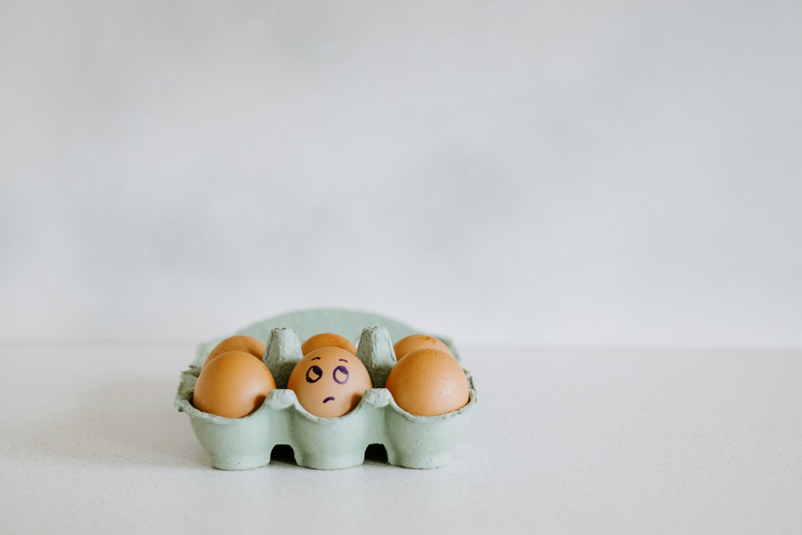 A carton of brown eggs sits on a white surface. A carton of brown eggs sits on a white surface; one egg has a worried face drawn on it with a marker, while the others are plain.
