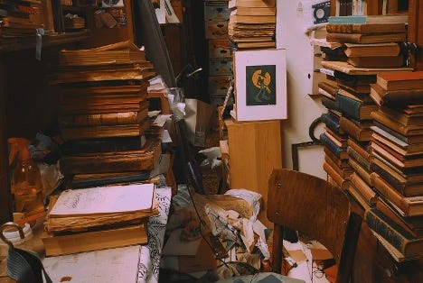 A cluttered room filled with tall stacks of books and papers A cluttered room filled with tall stacks of books and papers, wooden chairs, scattered objects, and some framed artwork leaning against the wall. The space appears crowded and disorganized, resembling a used bookstore or study.