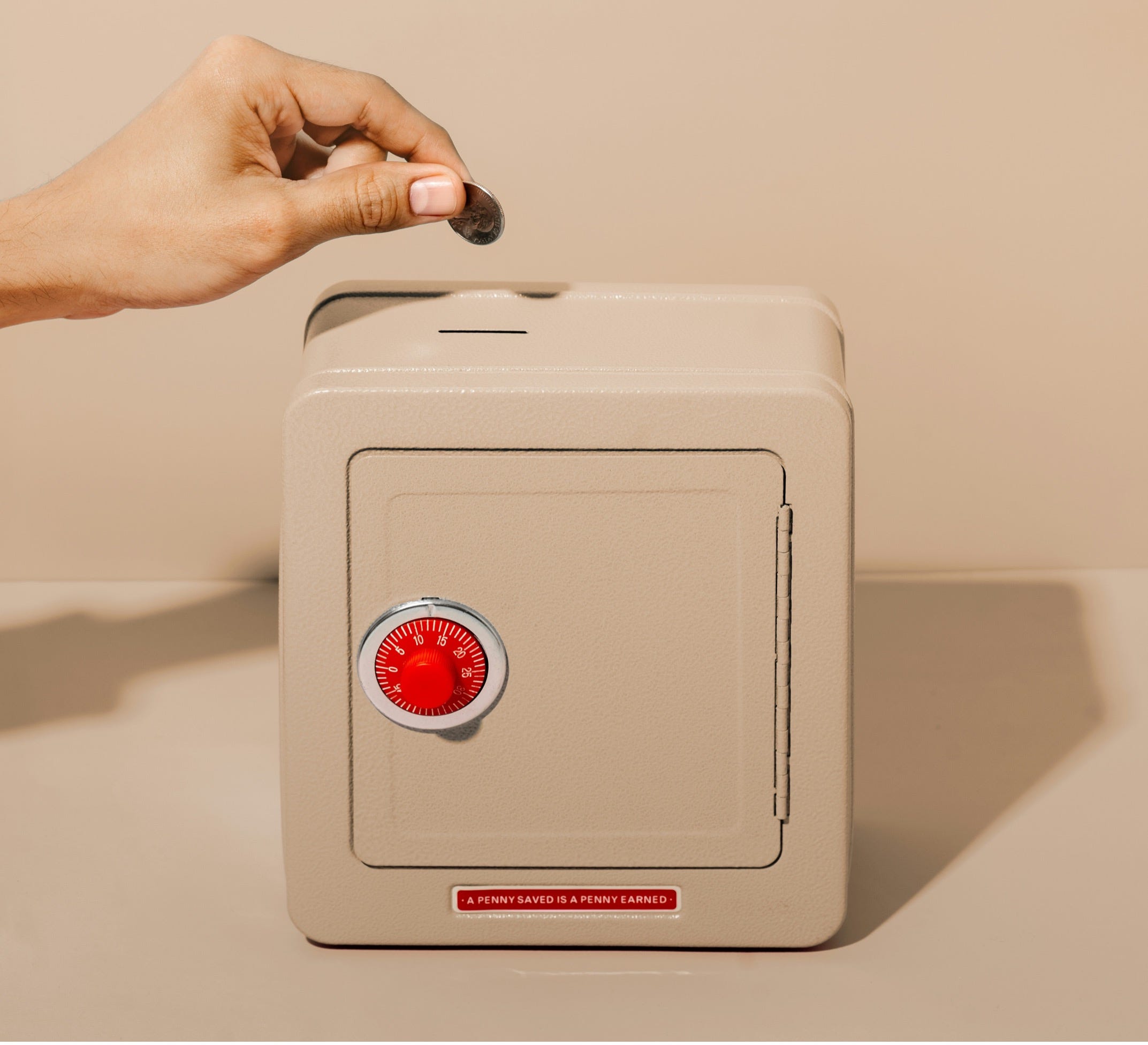 A hand drops a coin into a beige safe with a red combination lock A hand drops a coin into a beige safe with a red combination lock and a small label that reads, A penny saved is a penny earned, against a neutral background.