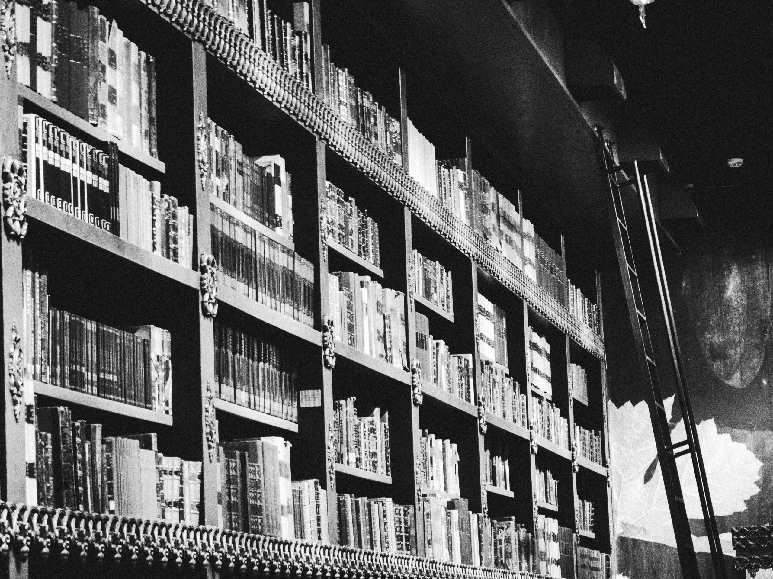 Black and white photo of tall, ornate bookshelves Black and white photo of tall, ornate bookshelves filled with books, extending upward and across the frame, with a ladder leaning against the shelves on the right side.