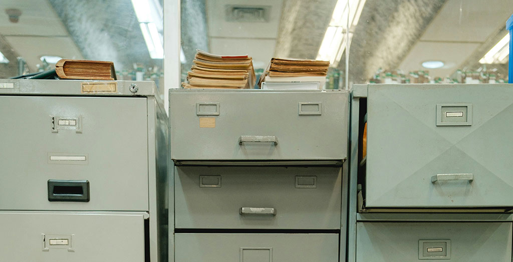 Three gray metal filing cabinets with drawers, some slightly open, stand side by side. Stacks of folders and papers rest on top—a look inside the records often managed by Public Library Boards. The background shows fluorescent lights and more cabinets. Three gray metal filing cabinets with drawers, some slightly open, stand side by side. Stacks of folders and papers rest on top—a look inside the records often managed by Public Library Boards. The background shows fluorescent lights and more cabinets.