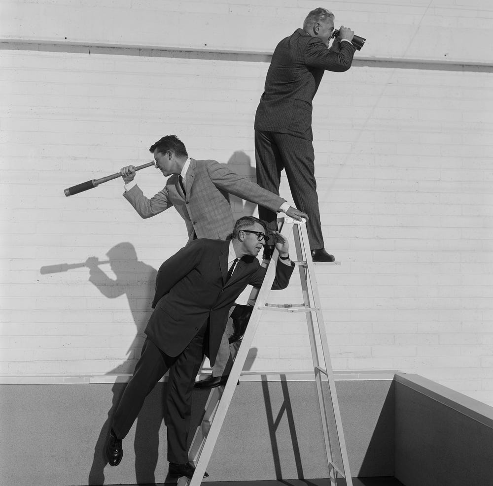 Three men in suits stand on a ladder against a wall, each looking in different directions using a telescope, binoculars, and periscope. The scene appears staged and humorous. Three men in suits stand on a ladder against a wall, each looking in different directions using a telescope, binoculars, and periscope. The scene appears staged and humorous.